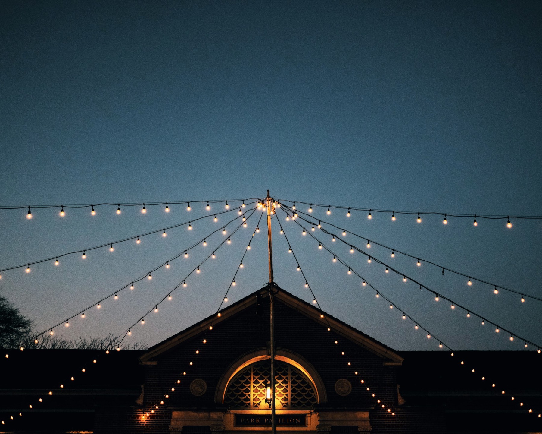 Strings of warm bulbs fanning out across a deep blue dusk sky above a courtyard pavilion.