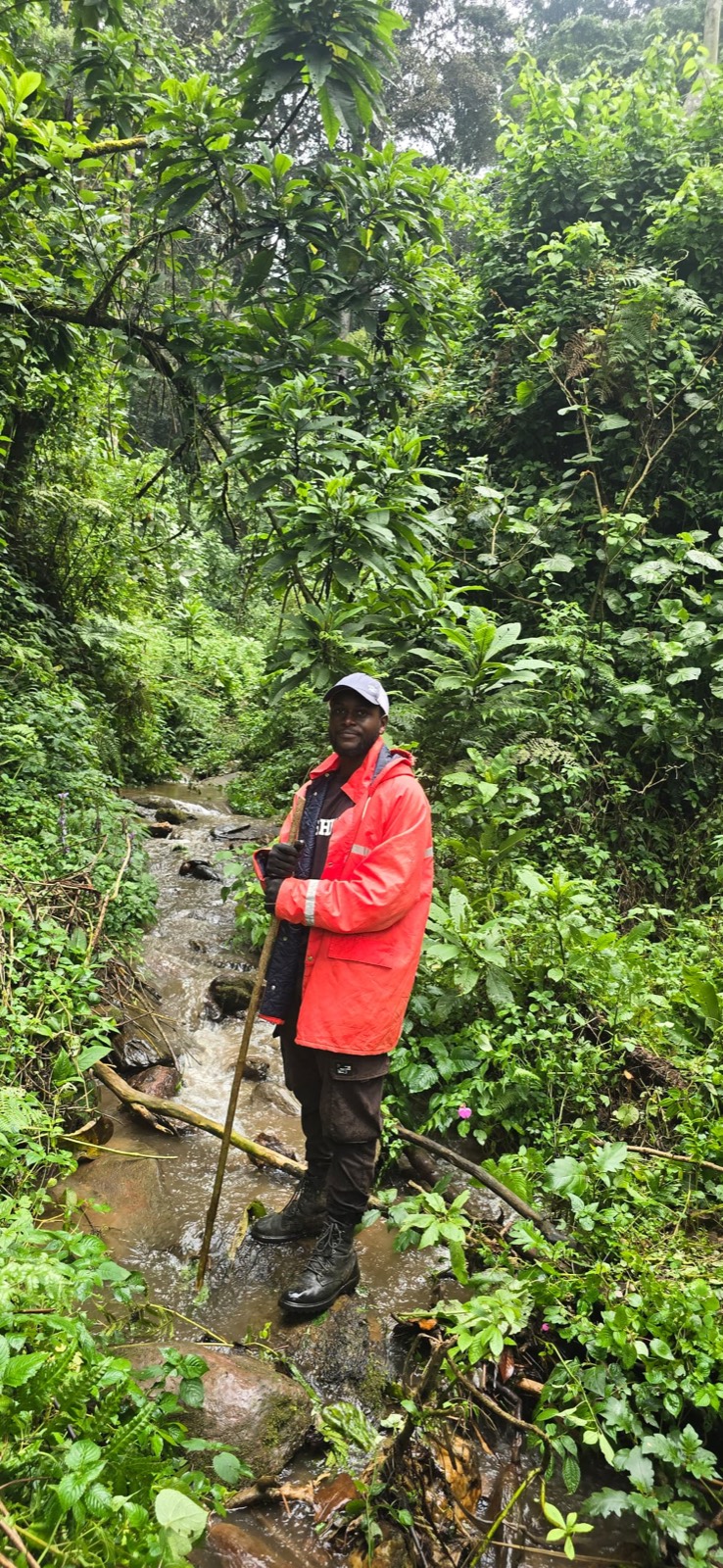 Yves Shema standing in a forest stream in heavy green undergrowth, wearing a red field jacket.